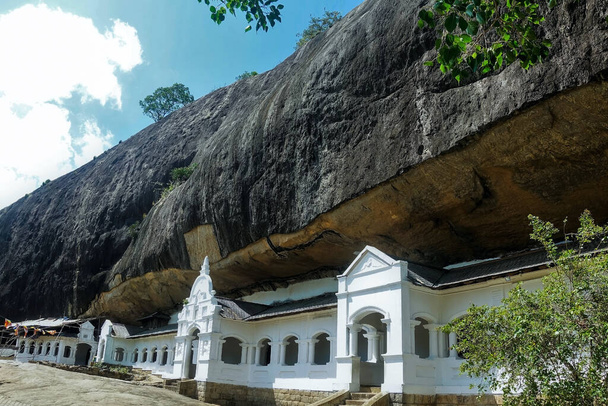 El antiguo templo cueva de Dambulla, Sri Lanka. Fachada de piedra blanca del edificio con ventanas y puertas talladas. Una enorme roca negra cuelga sobre el templo. Patrimonio de la UNESCO
. - Foto, imagen
