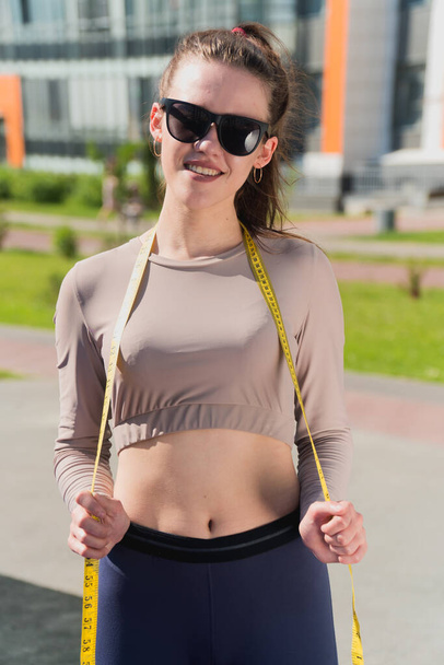 Happy slender girl holds a centimeter tape in her hands. The concept of improper weight loss woman fitness trainer in the park - Photo, Image
