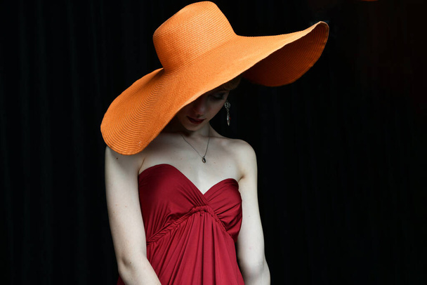 piękna dziewczyna w kapeluszu o szerokim rondzie i w burgundowej sukni wieczorowej pozująca na czarnym tle - Zdjęcie, obraz