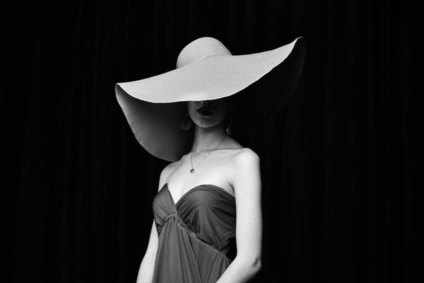 mooi meisje in een brede rand hoed en in een bordeaux avondjurk poseren op een zwarte achtergrond - Foto, afbeelding