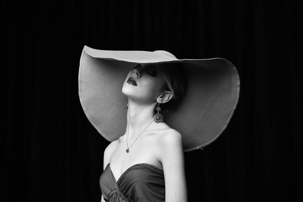beautiful girl in a wide-brimmed hat and in a burgundy evening dress posing on a black background - Photo, Image