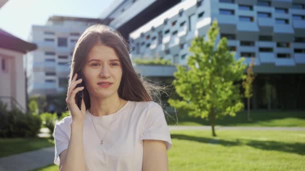 Gyönyörű lány beszél telefonon egy barátjával egy modern épület hátterében. Napsütéses nyári nap. - Felvétel, videó