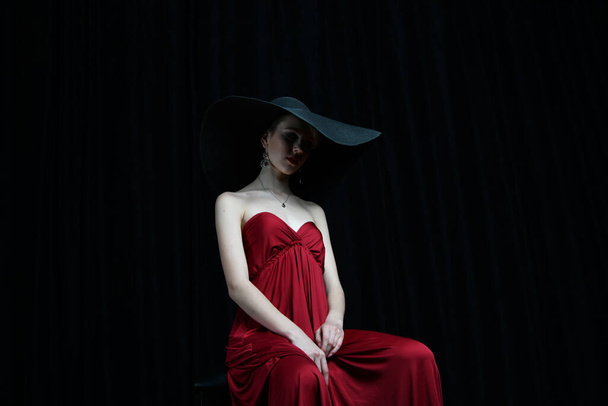 όμορφο κορίτσι σε ένα πλατύ καπέλο και σε ένα μπορντό βραδινό φόρεμα ποζάροντας σε μαύρο φόντο - Φωτογραφία, εικόνα