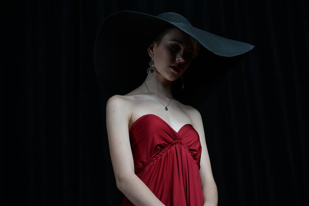 schönes Mädchen mit breitkrempigem Hut und weinrotem Abendkleid, das auf schwarzem Hintergrund posiert - Foto, Bild
