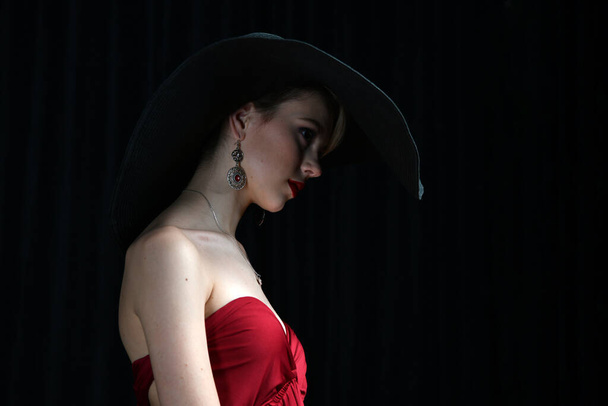 Geniş kenarlı şapkalı, bordo bir gece elbisesiyle siyah arka planda poz veren güzel bir kız. - Fotoğraf, Görsel
