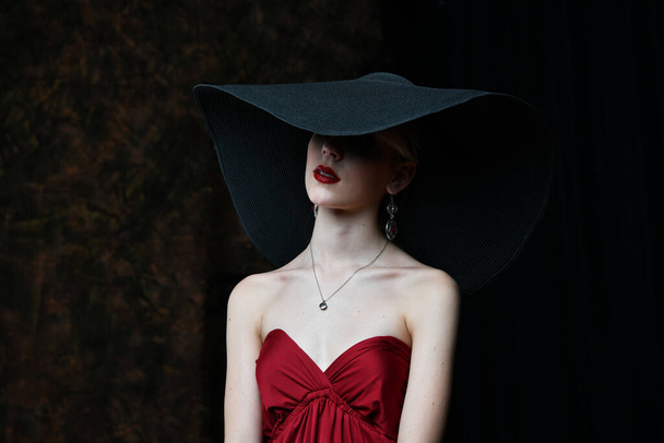 όμορφο κορίτσι σε ένα μπορντό καπέλο και σε ένα μπορντό βραδινό φόρεμα ποζάροντας σε μαύρο φόντο - Φωτογραφία, εικόνα