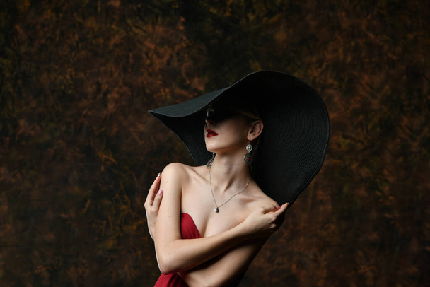 Siyah şapkalı ve bordo bir gece elbisesi giymiş güzel bir kız kendini kollarına sarmış. - Fotoğraf, Görsel