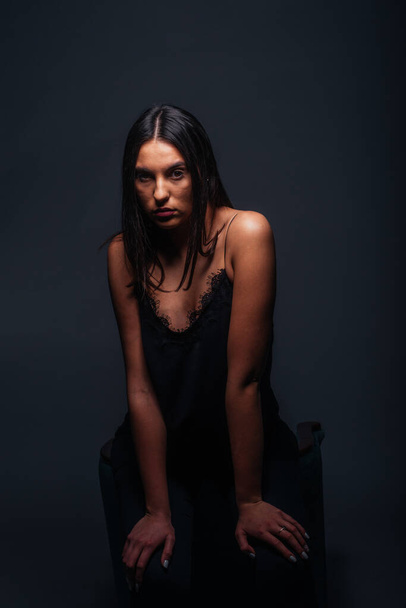 Πανέμορφο γυναικείο μοντέλο με σέξι μαύρο μπλουζάκι και μαύρο παντελόνι που ποζάρει μπροστά από μια καρέκλα μέσα σε ένα φωτογραφικό στούντιο - Φωτογραφία, εικόνα