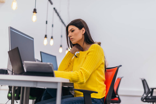 Chica hermosa concentrada con el pelo largo negro y blusa amarilla trabajando en un ordenador portátil en una oficina moderna. - Foto, imagen