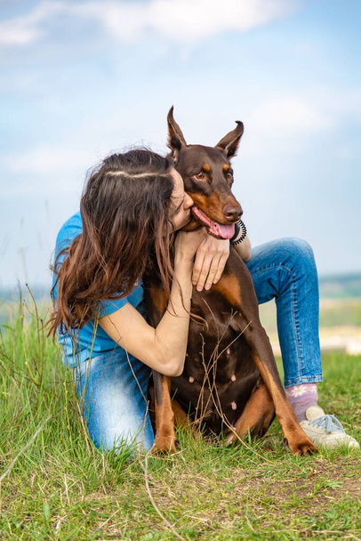 Una chica de fútbol azul y jeans se sienta abrazando y besando a un perro Doberman Dobermann marrón y bronceado sobre hierba verde en una colina contra el cielo. Orientación vertical
.  - Foto, imagen
