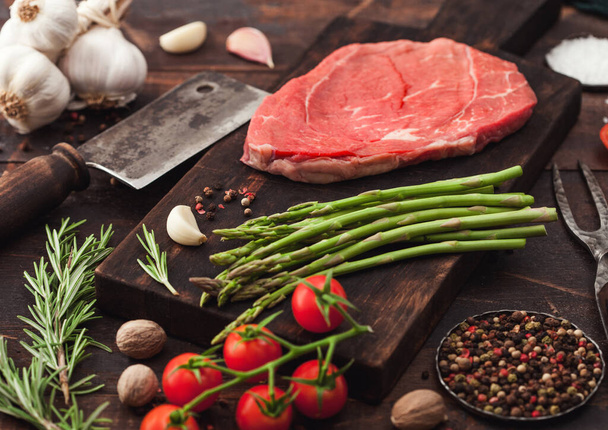 Slice of fresh raw barbeque braising beef steak on chopping board with asparagus and garlic with tomatoes and salt with pepper on wooden background with hatchet. - Zdjęcie, obraz