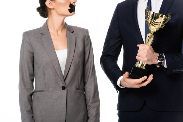 vista parcial del empresario sosteniendo trofeo de oro cerca de la mujer de negocios con cinta adhesiva en la boca y medalla aislada en blanco
 - Foto, Imagen