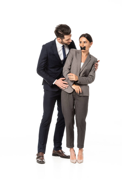 Geschäftsmann in offizieller Kleidung berührt Geschäftsfrau mit Klebeband auf Mund isoliert auf weißem, sexueller Belästigung Konzept   - Foto, Bild