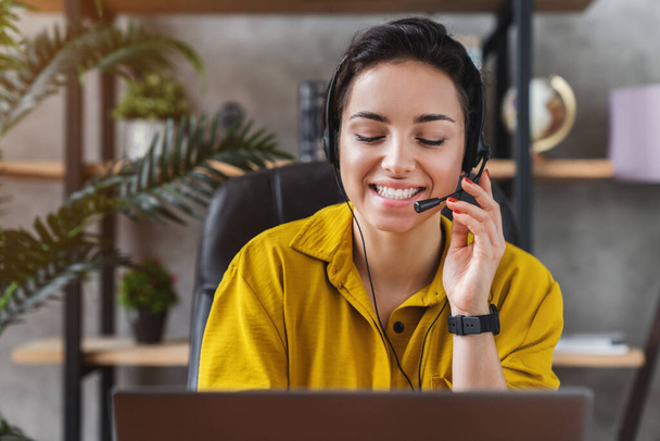 Smiling young female teleoperator working in home office with laptop and headset on - Foto, Bild