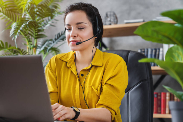Beautiful freelancer female talking in online video conference using headset with microphone and laptop in home office desktop - Fotoğraf, Görsel