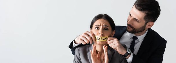 horizontální obraz obchodníka dotýkajícího se skotské pásky s nápisem sexismu na ústa vyděšené podnikatelky gestikulující izolovaně na bílém - Fotografie, Obrázek