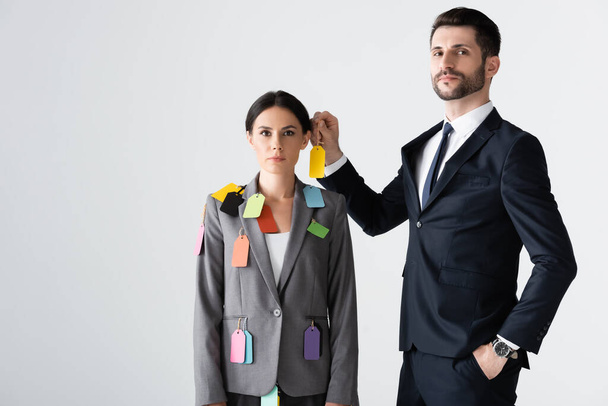 bearded businessman standing with hand in pocket and putting label on beautiful businesswoman isolated on white, sexism concept  - Fotó, kép