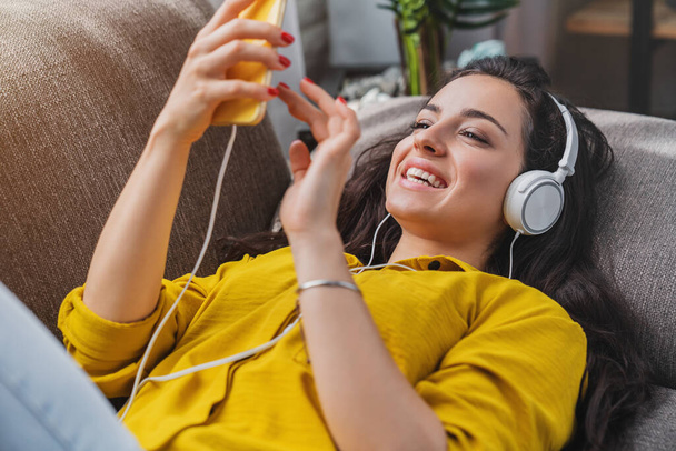 幸せな若い女性は自宅で音楽を聴くスマートフォンやヘッドフォンとソファに横たわっています - 写真・画像