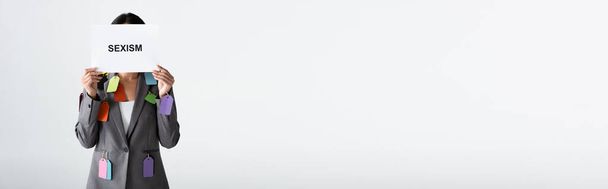 panoramatická plodina obchodnice s etiketami na skafandru s nápisem sexismu izolovaným na bílém - Fotografie, Obrázek