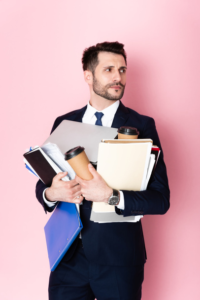 handsome businessman holding paper cups, laptop, folders and notebooks while looking away on pink  - Foto, imagen