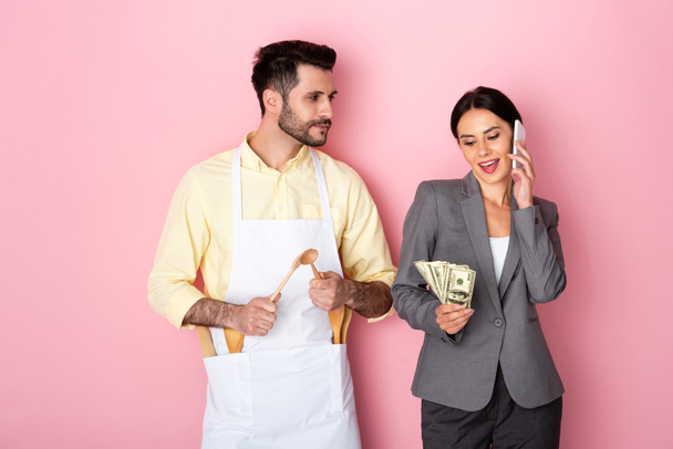 エプロンのハンサムな男が木製のスプーンを保持し、ピンクの上にスマートフォンで話してお金でビジネス女性を見て  - 写真・画像