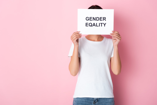 γυναίκα με λευκό t-shirt που καλύπτει το πρόσωπο με την ισότητα των φύλων γράμματα σε πλακάτ και στέκεται σε ροζ  - Φωτογραφία, εικόνα