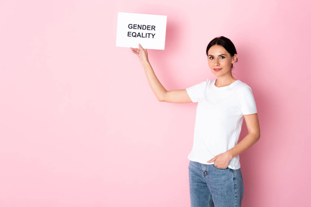 Beyaz tişörtlü mutlu bir kadın, elinde cinsiyet eşitliği yazılı bir pankart tutuyor ve pembe üzerinde el ele duruyor.  - Fotoğraf, Görsel