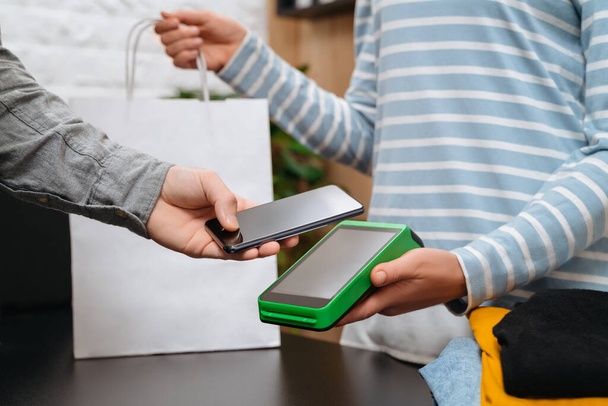 Απρόσκοπτη έννοια πληρωμής, αρσενικό πελάτη κρατώντας smartphone κοντά στην τεχνολογία NFC στον πάγκο, πελάτης κάνει τη συναλλαγή, να πληρώσει το λογαριασμό στο τερματικό rfid ταμίας στο κατάστημα ρούχων. Τεχνολογία NFC - Φωτογραφία, εικόνα