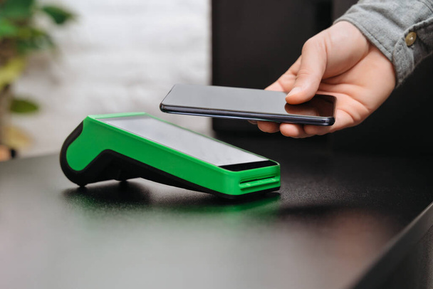 Ο άνθρωπος πληρώνει το λογαριασμό μέσω smartphone χρησιμοποιώντας την τεχνολογία NFC σε ένα κατάστημα. Άντρας πελάτης πληρώνει μέσω κινητού τηλεφώνου χρησιμοποιώντας τεχνολογία ανέπαφων συναλλαγών. Κοντινό πλάνο χέρι του ανθρώπου που κάνει την πληρωμή κινητό στο κατάστημα - Φωτογραφία, εικόνα