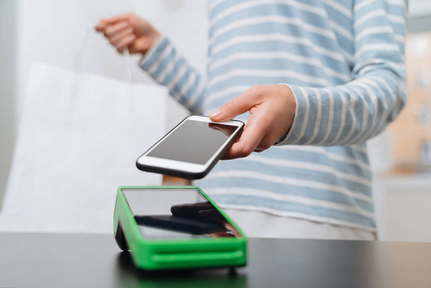 Naispuolinen asiakas maksaa ostosta älypuhelimella NFC-teknologian avulla. lähikuva naisen käsi maksaa kontaktittoman koneen tai NFC terminaali. Kontaktiton maksutapa - Valokuva, kuva