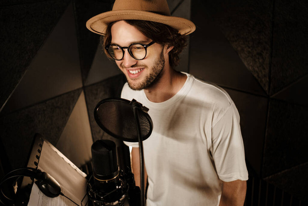 Chanteur masculin souriant et élégant enregistrant joyeusement une nouvelle chanson pour un album de musique dans un studio sonore moderne - Photo, image