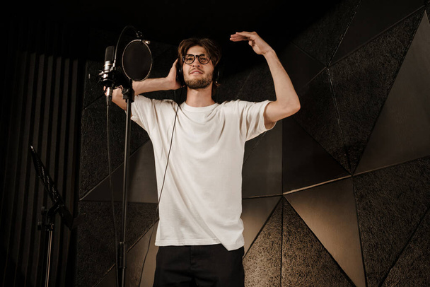 Młody atrakcyjny mężczyzna śpiewak sprawdza dźwięk w słuchawkach przygotowując nową piosenkę w nowoczesnym studio dźwiękowym  - Zdjęcie, obraz