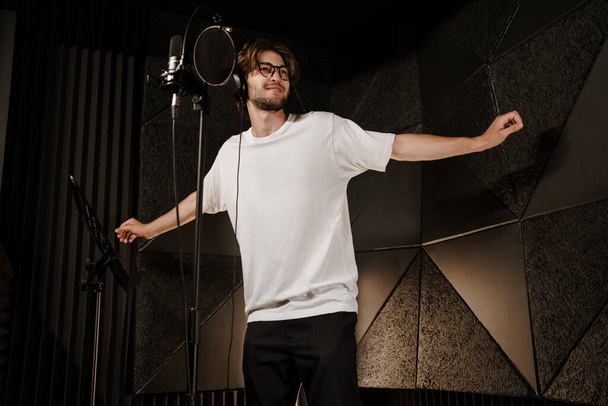 Joven cantante masculino atractivo en auriculares bailando durante la grabación de una nueva canción en el estudio de sonido moderno. Joven músico cantando en micrófono
 - Foto, imagen