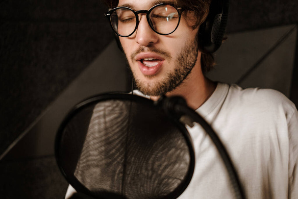 Detailní záběr pohledný mužský hudebník ve sluchátkách smyslně zpívat v mikrofonu v nahrávacím studiu  - Fotografie, Obrázek