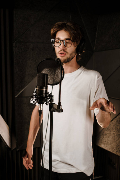 Genç, çekici, kulaklıklı erkek müzisyen modern ses stüdyosunda yeni bir müzik albümü için dikkatle şarkı kaydediyor.  - Fotoğraf, Görsel