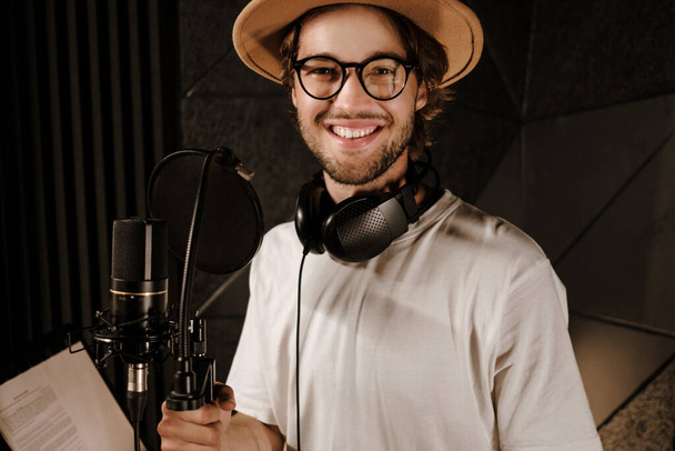 Ritratto di bel cantante maschio allegro che guarda felicemente in macchina fotografica durante il lavoro nel moderno studio di registrazione  - Foto, immagini