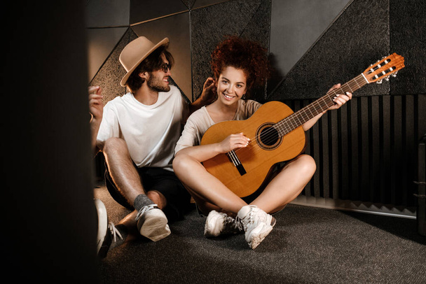 Mladý atraktivní muž a žena šťastně hraje novou píseň na kytaru sedí na podlaze v moderním nahrávacím studiu - Fotografie, Obrázek