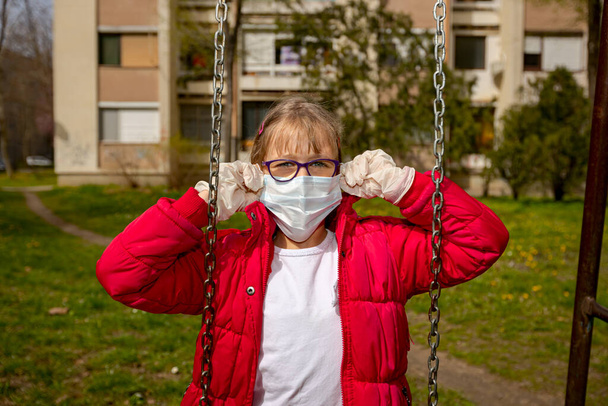 Tıbbi maskeli ve eldivenli küçük kız COVID-19 virüsünden korunmak için tek başına sallanıyor..  - Fotoğraf, Görsel