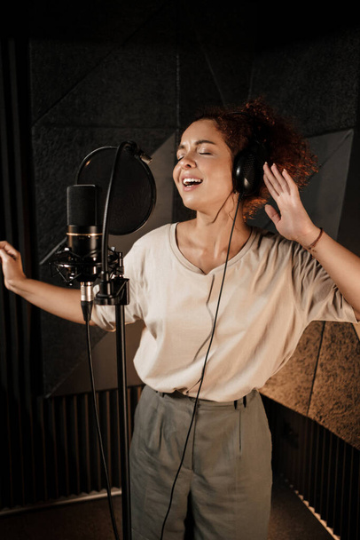 Attractive female musician in headphones emotionally singing in sound recording studio - Photo, Image