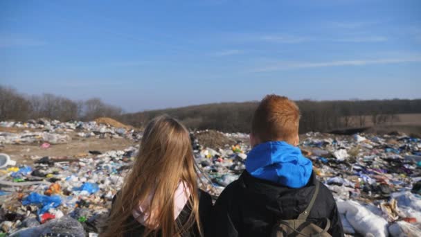 Achteraanzicht van jong meisje en jongen staat tegen de wazige achtergrond van vuile vuilnisbelt. Kleine kinderen kijken naar vuilnis op het autokerkhof. Begrip milieuvervuilingsprobleem. Sluiten. - Video