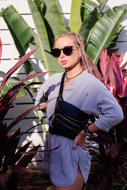 Young europian stylish woman with pink purple braids in gray long shirt, accessories, sunglasses, black waist bag trendy fashion watch clock on hand, on tropical background and white wooden wall. - Foto, imagen