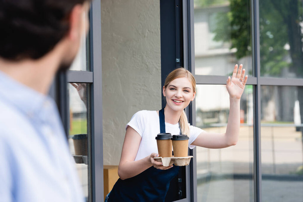 Selective focus of smiling waitress holding paper cups and waving hand at man near cafe  - Foto, afbeelding