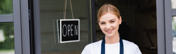 Panoramic shot of smiling waitress looking at camera near signboard with open lettering on door of cafe - Photo, Image