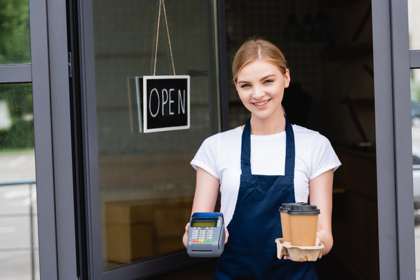 Camarera sonriente sosteniendo terminal de pago y café para ir cerca del letrero con letras abiertas en la puerta de la cafetería
  - Foto, Imagen