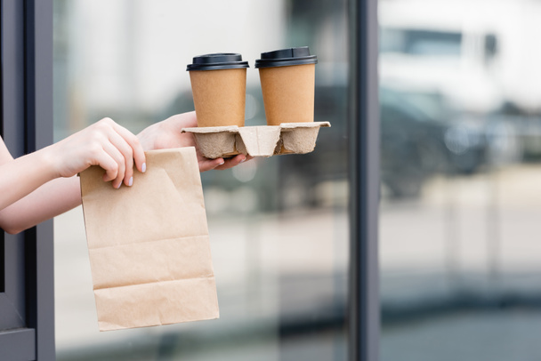 Vista ritagliata della cameriera con sacchetto di carta e caffè per andare vicino al caffè sulla strada urbana  - Foto, immagini