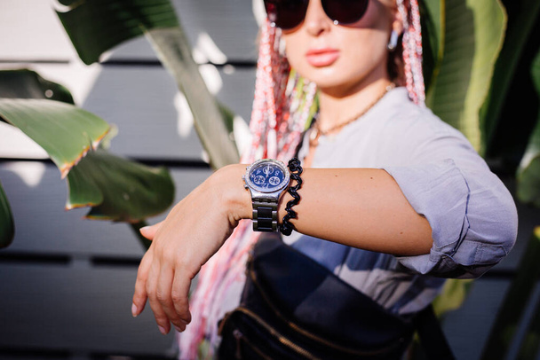 Mladá evropsky stylová žena s růžovými fialovými copánky v šedé dlouhé košili, doplňky, sluneční brýle, černý pas taška módní hodinky po ruce, na tropickém pozadí a bílé dřevěné zdi. - Fotografie, Obrázek
