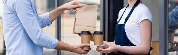 Panoramic crop of waitress giving disposable cups and paper bag to customer near cafe on urban street - Photo, Image