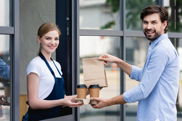 Camarera sonriente y cliente sosteniendo café para llevar y bolsa de papel mientras sonríe a la cámara cerca de la cafetería en la calle urbana
  - Foto, imagen