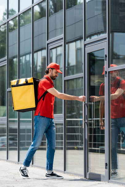 Delivery man in uniform and thermo backpack opening door of building on urban street  - Foto, imagen