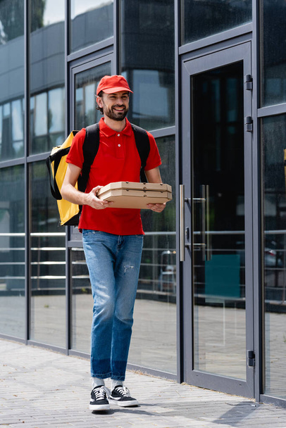 Smiling courier with thermo backpack holding pizza boxes while walking near building on urban street  - Foto, afbeelding